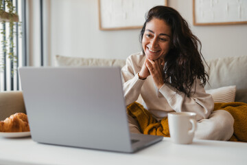 Smiling woman sitting comfortably on a sofa at home, looking at her laptop with a warm and positive...