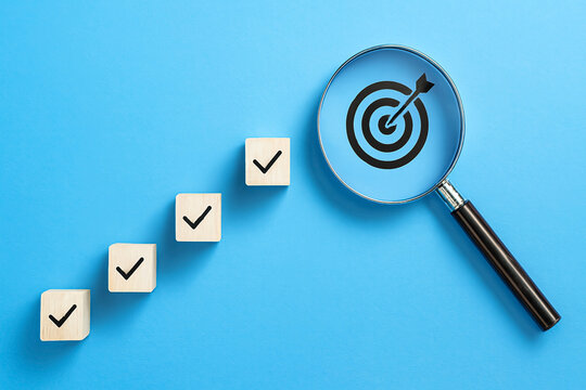 Wooden blocks with checkmarks ascending diagonally toward magnifying glass highlighting target with arrow bullseye on blue background. Conceptual 3D rendering symbolizing focus, progress, and goal ach