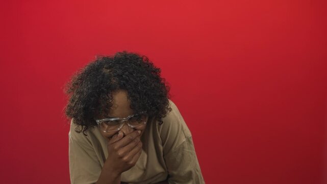 Young african american woman wearing glasses covers mouth with hand in red studio; amusement shyness warmth.