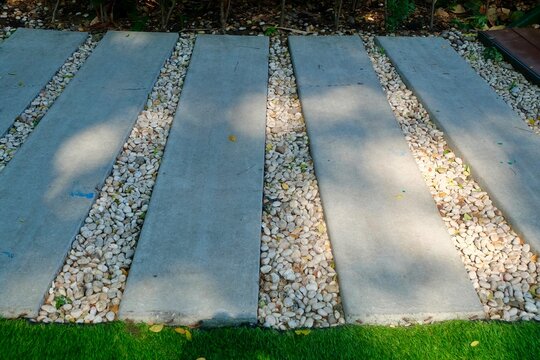 A contemporary garden path features parallel rectangular concrete slabs separated by channels filled with white pebbles, bordered by artificial green grass, under dappled sunlight.