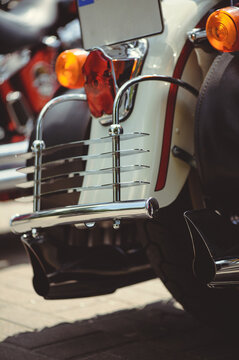 Detailed shot of the rear section of a classic motorcycle, featuring a white fender with red pinstripes, a polished chrome protective rack, a red tail light, and orange circular turn signals.