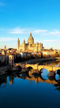 A breathtaking view of Salamanca's historic cathedral and Roman bridge reflected in the Tormes River during a serene golden hour sunset.