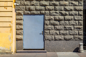 simple blue door stands embedded in a rough-hewn stone wall, flanked by a weathered yellow facade. The scene conveys a sense of quiet urban durability, with a security camera overhead adding a modern 