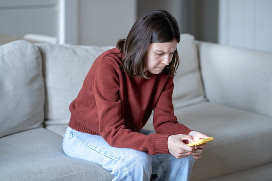 Stressed woman frowning while checking smartphone, scrolling social media sits on sofa at home. Tense nervous young female reading phone messages with worried expression, feeling anxiety, uncertainty
