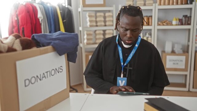 Man tapping tablet and sorting clothes donations at a donations desk inside a volunteer center building with labeled boxes and shelves; compassion service.