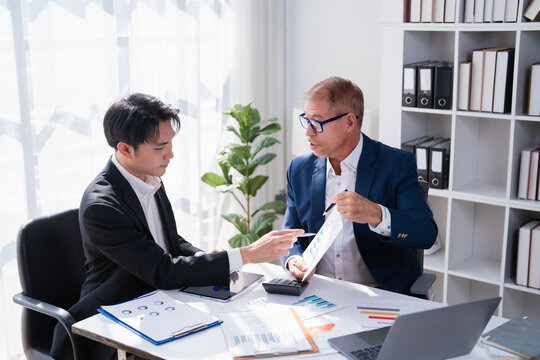 Two businessmen collaborate in a modern office, reviewing financial reports, charts and a laptop as they discuss strategy, analyze data and plan corporate growth during a meeting