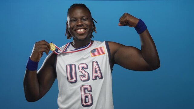 Man wearing usa jersey with flag patch holding a gold medal by the ribbon and flexing bicep while smiling in a bright blue studio with wristbands; victory pride celebration.