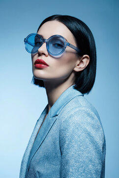 Stylish woman with blue sunglasses and shimmering jacket posing against a soft blue background.