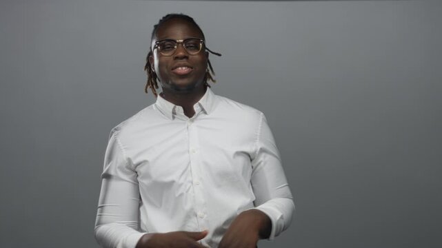 Young african american man with glasses and dreadlocks gesturing open hands in a gray studio wearing white shirt; confidence.
