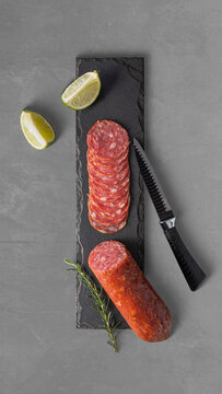 Salame artesanal fatiado sobre t&aacute;bua de ard&oacute;sia, com faca serrilhada, alecrim e lim&atilde;o. Composi&ccedil;&atilde;o flat lay minimalista, ilumina&ccedil;&atilde;o de est&uacute;dio e fundo cinza, ideal para conceitos de gastronomia gourmet
