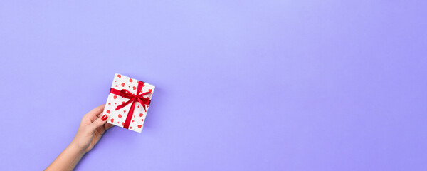 Obraz premium Woman hands give wrapped valentine or other holiday handmade present in paper with red ribbon. Present box, red heart decoration of gift on violet table, top view with copy space for you design
