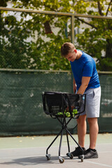 Fototapeta premium Young man organizing tennis balls in a basket on a sunny day at an outdoor court while preparing for practice session