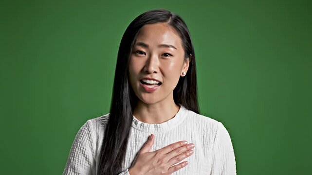 Asian woman taking an oath or promise with her hand on her heart in a studio with a green background