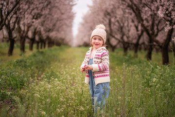 Young girl standing in green grass under pink blossoming trees, smiling and holding a tiny flower on a sunny spring day, capturing innocence, joy and playful outdoor freedom