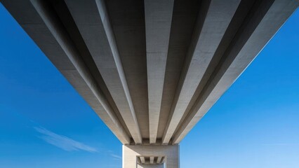 Naklejka premium Underneath a concrete highway bridge, parallel beams stretch toward a square pillar, set against a bright blue sky. Concept Industrial Architecture, Underpass Geometry, Parallel Beams, Square Pillar