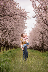 Mother tenderly embracing her baby daughter, standing together in a lush green field surrounded by rows of beautiful pink cherry blossom trees overhead, symbolizing family love and spring happiness