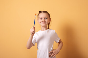 Cheerful young girl in a white t-shirt holding a paintbrush against a solid pastel orange background, ideal for creative and educational concepts.