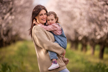 Mother smiling and embracing her young daughter, both enjoying a warm family moment together outdoors in a beautiful blooming almond tree orchard filled with white and pink flowers