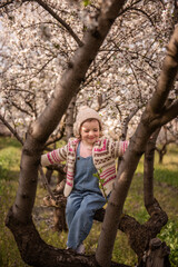 Young girl smiling and sitting on a tree branch, playing outdoors in a blossoming almond orchard, embodying childhood happiness and freedom in nature during springtime