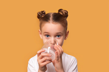 Young girl with brown hair in buns drinks water from a glass, looking at camera, on solid orange background. Bright, healthy lifestyle concept.
