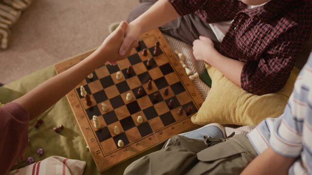 High angle cropped shot of unrecognizable kid winning chess game with checkmate and shaking hands with his rival, sitting on couch and playing board game at home