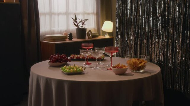 No people view of round table covered with tablecloth featuring cheese, grapes and skewer canapes on plates, chips in bowl and six empty glass goblets