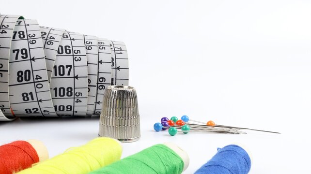 Spools of colored threads lined up in a row, a tape measure, pins and needles, and a steel thimble are placed on a white surface. Sewing and haberdashery equipment. Close-up.