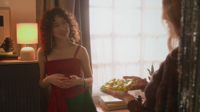 Young woman holding canapes with cheese and green grapes on plate and speaking with curly-haired girl friend, putting it on round table