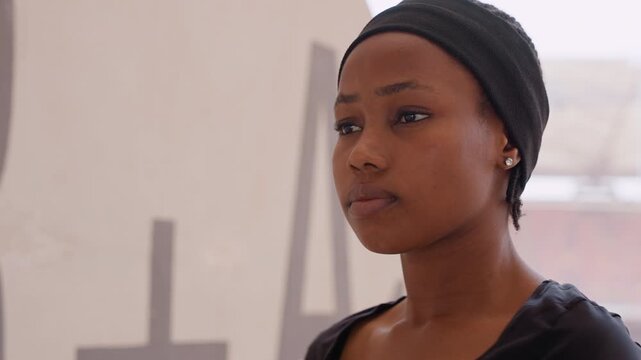 young black woman in studio soft natural light highlights headwrap and profile, contemplative expressions shifting from thoughtful to resolved as she poses as artist, model and student beside canvas