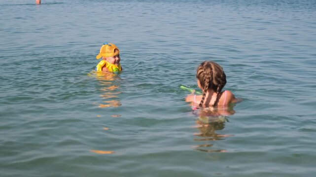 A little boy in a life jacket swims in the sea with a girl. The brother and sister splash and play in the seawater.