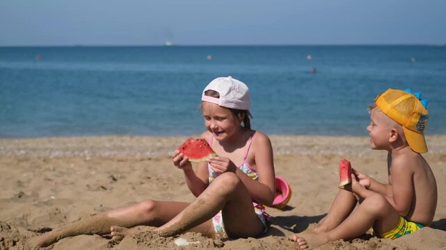 Children are sitting by the sea and eating watermelon. A sister and brother are sitting on the beach and having fun.