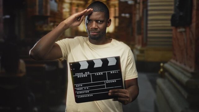Man salutes with right hand while holding a clapperboard in a narrow urban street with shuttered storefronts and stone steps; confidence cinema.