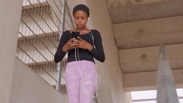 black woman rehearsing speech on stairwell, scrolling phone for notes while practicing gestures, wired earphones, confident posture and expressive hand movement against raw concrete and urban