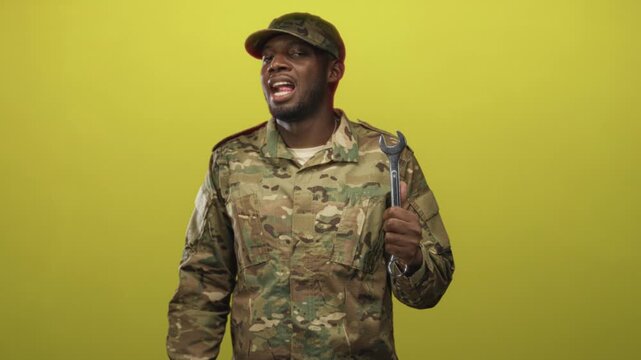 Young black man soldier in camouflage jacket and cap holding large wrench, sticking tongue out in studio; playful pride.