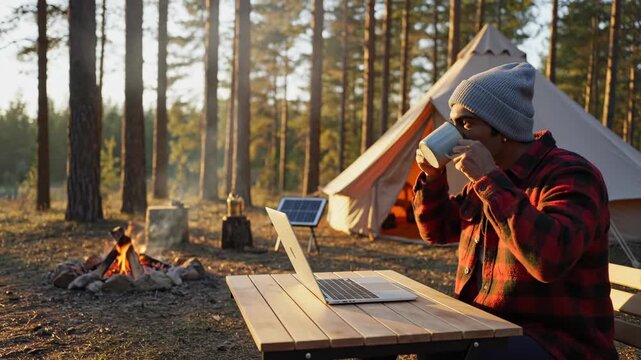 Man working on laptop at forest campsite. Remote work and digital nomad lifestyle. Golden hour sunlight