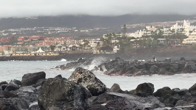 Waves crashing on rocky coastline near seaside resort town under cloudy sky