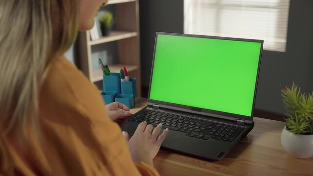 Woman focused on typing on a laptop with green chroma key screen at a home office desk, remote work or online learning setup with customizable display and copy space