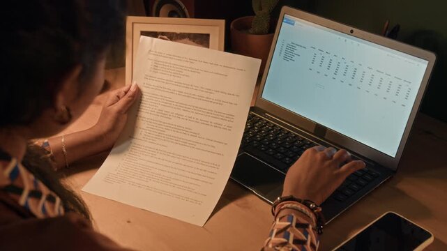 Over shoulder shot of young adult female remote employee filling out digital table entering data on laptop keyboard while analyzing document at home office desk