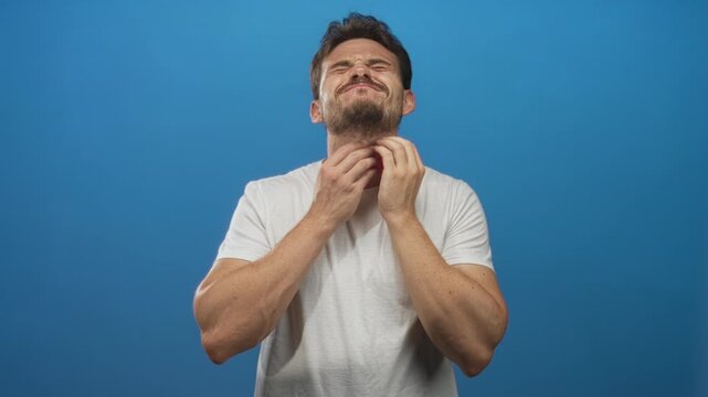 Young hispanic man in white t shirt clutching throat in studio with bright blue background; sore illness pain.