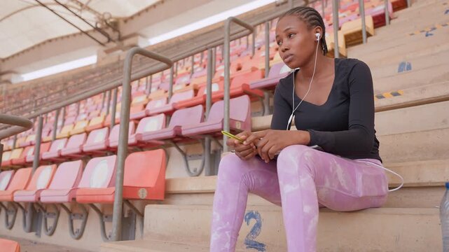 black teen scrolling phone on stadium bleachers, casual portrait showing young black woman with earphones and pink pants, hands holding smartphone, relaxed posture, covered roof and empty rows, subtle