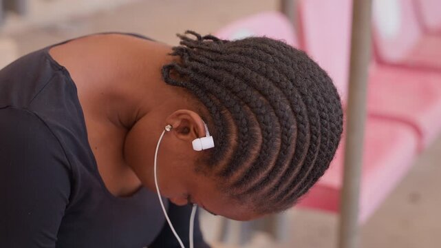 black teen commuter bowed head earbuds showing quiet fatigue and reflection on pink transit seats closeup of cornrow hairstyle and hand touching forehead, candid portrait capturing waiting, mood,
