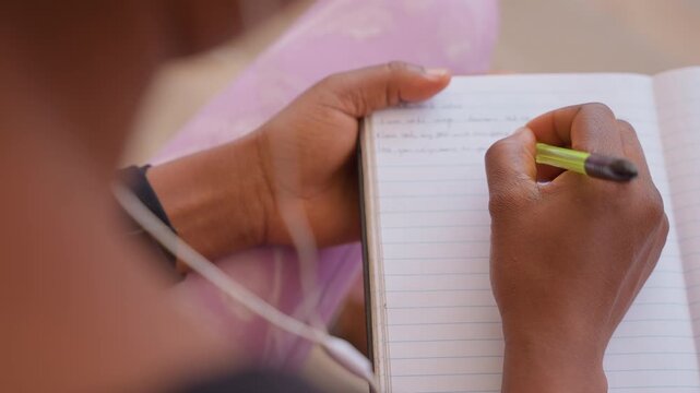 student writing in lined notebook closeup of hands holding green pencil, visible earphone cable and soft pink fabric backdrop, focused study and neat handwriting capturing reflective planning