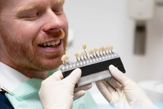 Dentist with gloves holding a tooth shade guide near a smiling patient's mouth