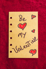 Be My Valentine, written on a piece of paper, on a red textile background.