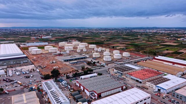 Flight over the warehouses approaching the oil refinery factory. Big and small tanks for fuel storage are in the scenery. Agricultural fields and cityscape at backdrop.