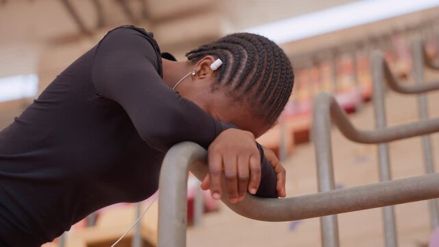black woman runner catching breath on railing, braided cornrows and earbuds, head bowed over metal bar, emotional exhaustion and solitude in empty stands, musicled recovery pause and inner resilience