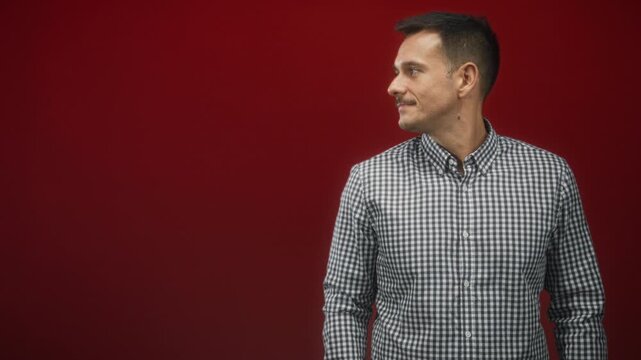Young man with moustache looks right, face turned, wearing checkered shirt in studio red backdrop; quiet confidence.
