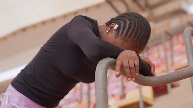 black performer pausing on railing stadium, young black woman with braided hair and earbuds taking breath between takes, empty bleachers and high canopy overhead, rehearsal atmosphere mixed with quiet