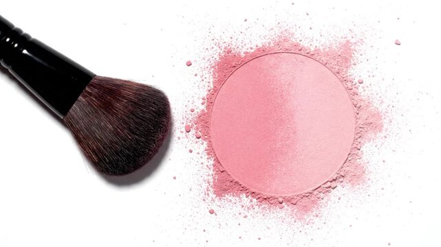 A makeup brush next to a pink blush compact with scattered powder on a white background.
