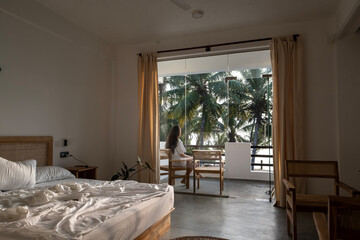 Obraz premium A young woman with long dark hair and a white T-shirt walks out onto the balcony from her luxurious bed after waking up in her hotel room. Wake-up time in a hotel room in Sri Lanka on vacation.